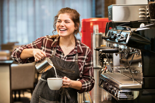 Female barista preparing coffee drink for customer at small Cafe, pouring milk while turning and smiling at customer        