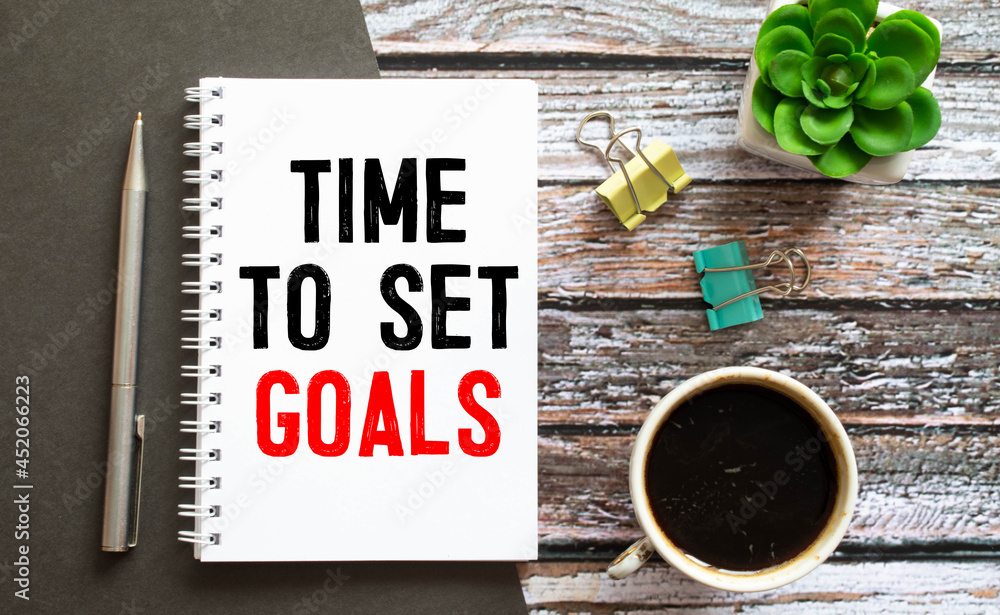 Poster time to set goals is written in a white notepad near a white alarm clock on a black background