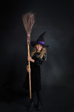 Little girl on a dark background in a witch costume and a wide-brimmed hat with a broom for Halloween.