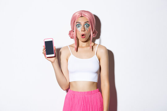 Image of surprised girl looking in awe while showing smartphone screen, dressed in pink wig and party outfit, standing over white background