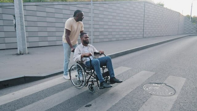 African American adult man crossing road at crosswalk pushing young bearded guy on wheelchair. Elderly father caring for non-walking, paralyzed son injured in accident. Accessible environment concept