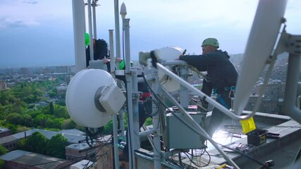 Workers servicing cellular antenna in front of city . Contractors working on a telecommunications mast at height . Telecommunication technician working on top of cellular antenna. The cell tower - Powered by Adobe