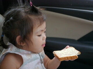A baby girl, 2 years old, learning to eat, by herself, a sheet of white bread with sweetened condensed milk on top in a car during a trip