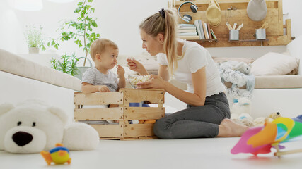 Happy and smiling mom with the beautiful baby child inside a wooden box, eating pop corn sitting at...
