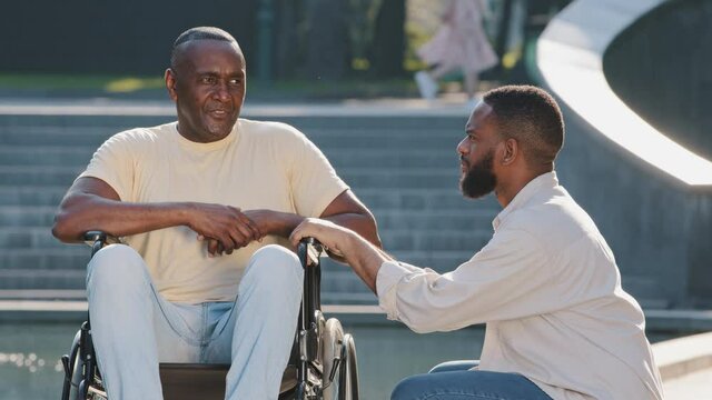 Young African American guy squatting down talking to elderly father in wheelchair. Healthy black man friendly with disabled person. Patient care, equal opportunity, inclusive environment concept