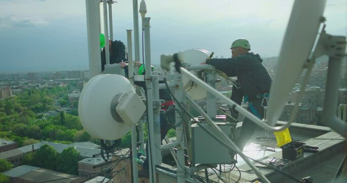 Workers Servicing Cellular Antenna In Front Of City . Contractors Working On A Telecommunications Mast At Height .  Telecommunication Technician Working On Top Of Cellular Antenna. The Cell Tower.