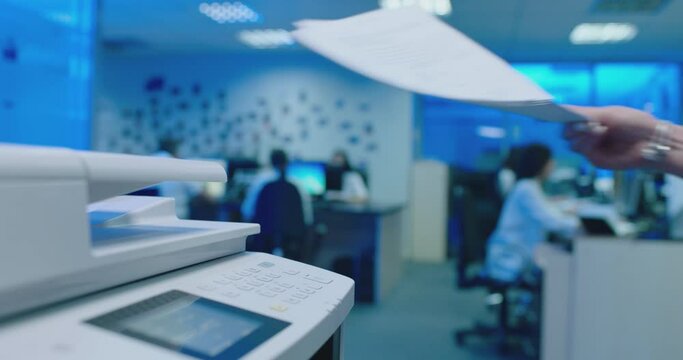 Woman using printer or scanner in office . Office worker use print machine on blurred office background with workers and computers . Hand take papers and press buttons on multifunctional copier .