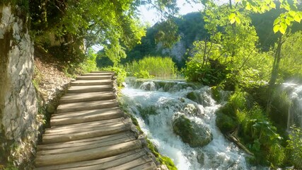 LENS FLARE: Wooden steps lead up to the top of a small sunlit waterfall in Plitvice. Empty boardwalk leads past a beautiful rushing crystal clear waterfall. Plitvice national park on a sunny day.