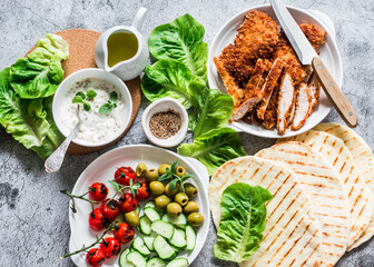 Ingredients for cooking greek gyros - chicken schnitzel, lettuce, tortillas, greek yogurt sauce, tomatoes, olives on a gray background, top view