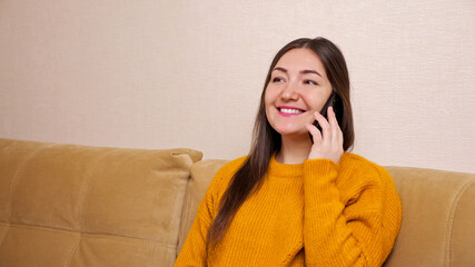 Young woman with long loose hair talks on black smartphone and smiles cheerfully sitting on brown...
