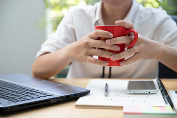 The hand of a businesswoman is lifting a cup of coffee on the table with laptop computer on...