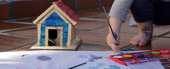 Mão de uma menina, segurando um pincel e pintando com aquarela em papel e em brinquedo de madeira