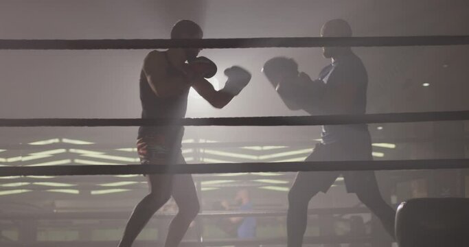 Male boxer training practicing with his trainer in low light gym . Silhouette on dark background. Boxer training in ring . Cinematic slow motion shot of professional young man punching with instructor