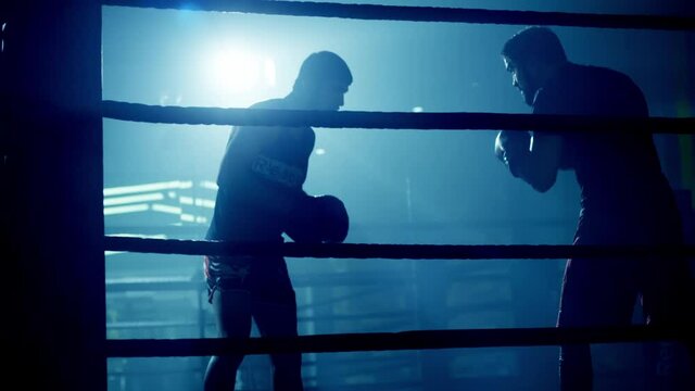 Male boxer training practicing with his trainer in low light gym . Silhouette on dark background. Boxer training in ring . Cinematic slow motion shot of professional young man punching with instructor
