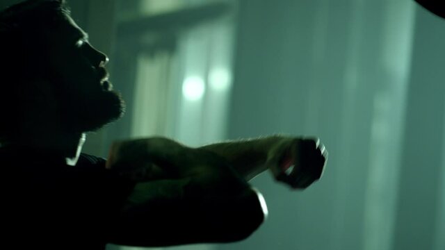 Young professional boxer boxing speed punching bag . Side view of young boxer practicing hand speed drill in slow motion using small punching bag in boxing gym .  Sportsman boxing in smoky studio .