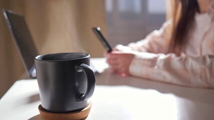 Close up of mug with hot drink on table against background of unrecognizable woman with phone and laptop zoom out
