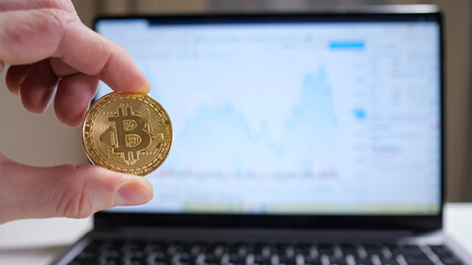 Male hand twirls bitcoin on the background of the laptop, showing the graph.