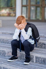A schoolboy with red hair sits on the stairs by the school, burying his face with his hands and...