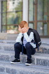 A schoolboy with red hair sits on the stairs by the school, burying his face with his hands and...