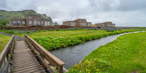 Fototapeta na wymiar Panoramic view over new modern houses, yellow meadow flowers, small creek and bridge in Vik town, South Iceland, at summer.