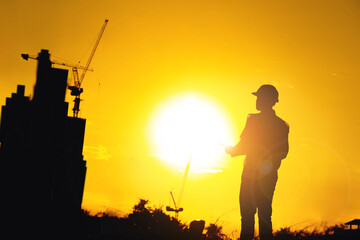 Silhouette of an engineer holding a yellow safety helmet with a tall apartment building and a construction crane on the background of the evening sunset. evening sky
