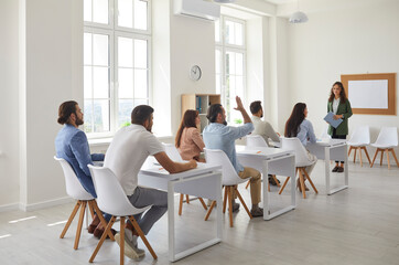 Group of workers sitting at tables in the classroom in a lesson with a professional business coach. Adult student raises his hand to ask a question to the speaker. Concept of professional development.