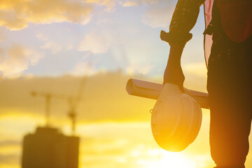 Silhouette of an engineer holding a yellow safety helmet with a tall apartment building and a construction crane on the background of the evening sunset. evening sky
