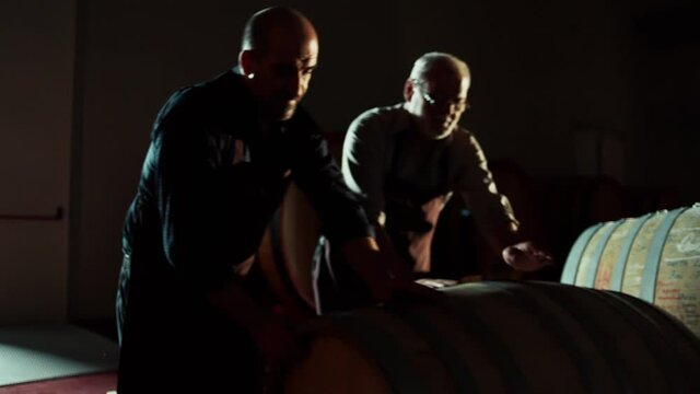 Worker in wine , whiskey or brandy warehouse sorting and rotating barrel . Two winemakers in vintage , traditional wine factory rolls barrel . Shot on ARRI ALEXA Cinema Camera in Slow Motion