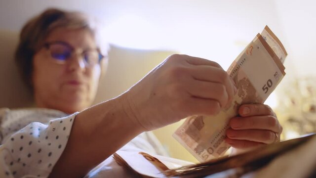 Concentrated middle-aged woman in eyeglasses lying in bed and counting euro paper currency from two packs, sad woman counting her monthly income, saving money for paying bills and property loan