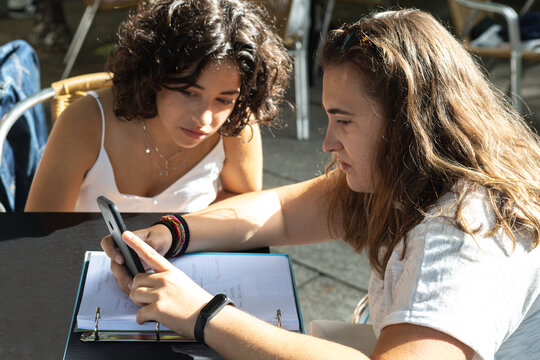 two young women studying and looking at their cell phones enjoying a sunny day at the table of a cafeteria terrace - Powered by Adobe