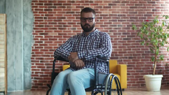 Thoughtful, bearded, disabled man with glasses is sitting in wheelchair, sad and depressed. Hipster disabled man sadly lowers his head, front view. Rehabilitation concept