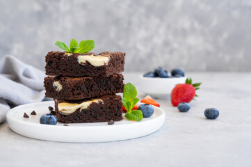 Delicious chocolate brownie cheesecake with fresh berries and mint on a white plate on a gray concrete background. Copy space.