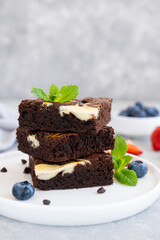 Delicious chocolate brownie cheesecake with fresh berries and mint on a white plate on a gray concrete background. Copy space.