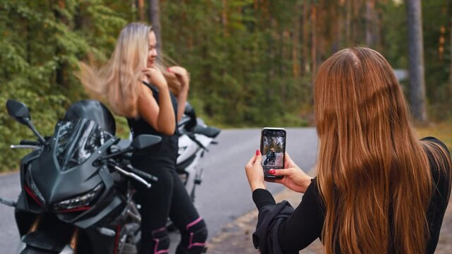 A woman with long hair taking a picture of her blonde girlfriend posing near the motorbike