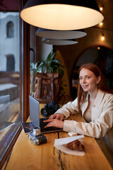 Smiling happy young female photographer sitting with laptop and camera at cafe. Concept of modern technology and photoartist. Side view portrait. Caucasian redhead female on freelance