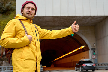 road trip, travel, gesture and people concept. caucasian young man hitchhiking and stopping car with thumbs up gesture at countryside, in yellow coat