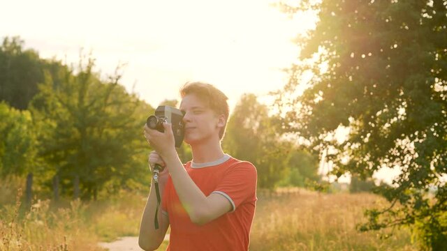 Close-up view of one young white handsome teenage boy using old vintage camera while standing outdoors in sunset scenic countryside landscape background