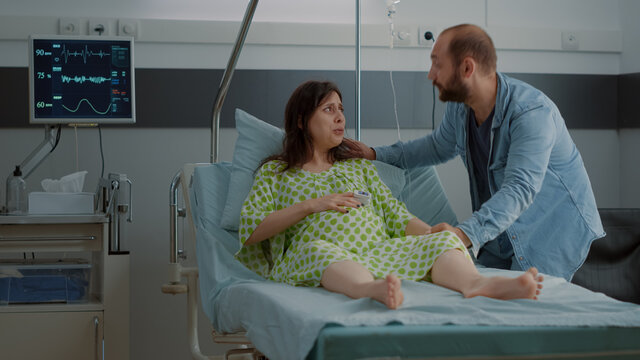 Young Woman Going Into Labor In Hospital Ward Sitting With Husband Holding Hand. Caucasian Patient Doing Breathing Exercices While Having Contractions Asking For Medical Assistance