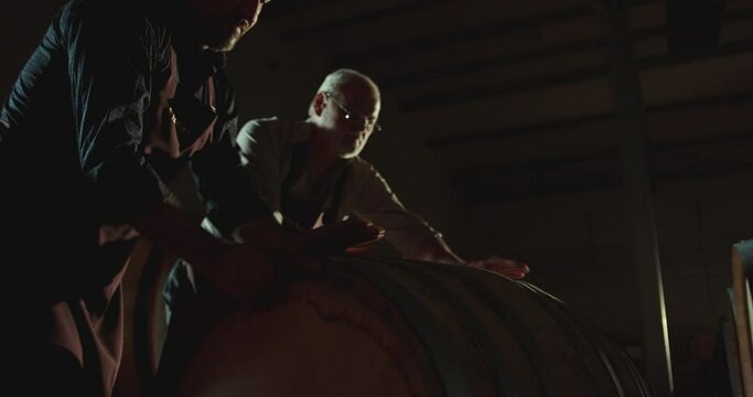 4k Worker in wine , whiskey or brandy warehouse sorting and rotating barrel . Two winemakers in vintage , traditional wine factory rolls barrel . Shot on ARRI ALEXA Cinema Camera in Slow Motion