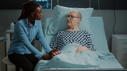 Sick elder patient laying in bed talks to african american visitor in hospital ward. Old man with...
