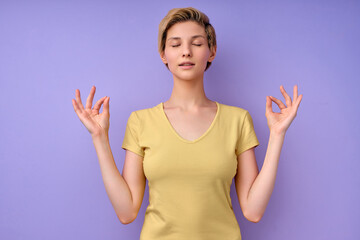 Young female in casual wear keep calm, meditating with eyes closed in studio on purple background, short-haired woman engaged in yoga, namaste. Nice lady of caucasian apperance copy space