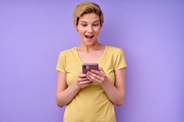 Amazed young woman in casual t-shirt, keeping mouth widely opened,looking at smartphone, caucasian beautiful model posing isolated over purple background, reading shocking news. copy space