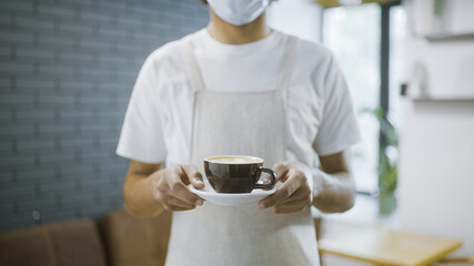 Fototapeta na wymiar Waiter in protective mask offering cup of coffee to guest, restaurant service