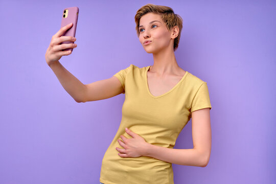 Gorgeous lady in casual outfit posing at camera taking selfie on smartphone, kissing. Side view portrait of woman with short hair taking picture on mobile phone, posing, isolated purple background