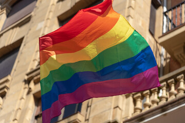 Una bandera arco iris, simbología lgtbi ondeando al viento con un edificio marrón detrás. - obrazy, fototapety, plakaty