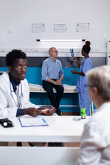 Fototapeta premium African american medic discussing with old woman sitting at office desk. Black nurse holding x ray scan explaining healthcare diagnosis to elderly man sitting on bed in medical cabinet