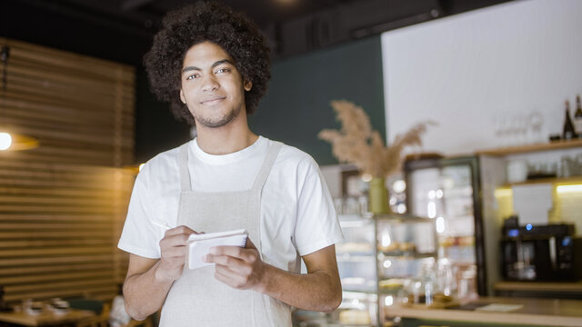 Coffee shop worker writing order in notebook, smiling at camera, part-time job