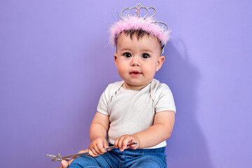 Pretty baby girl wearing princess crown on head, holding magic wand, caucasian child is looking at camera, pastel purple studio wall, copy space. happy childhood concept