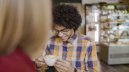 Happy mixed-race young man drinking coffee and talking to female friend, relax in cafe