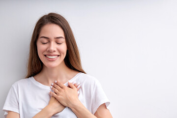 Pretty happy woman expressing gratitude, holding hands on chest, thankful for everything, with eyes closed, having nice smile, casually dressed. isolated on white studio background, copy space